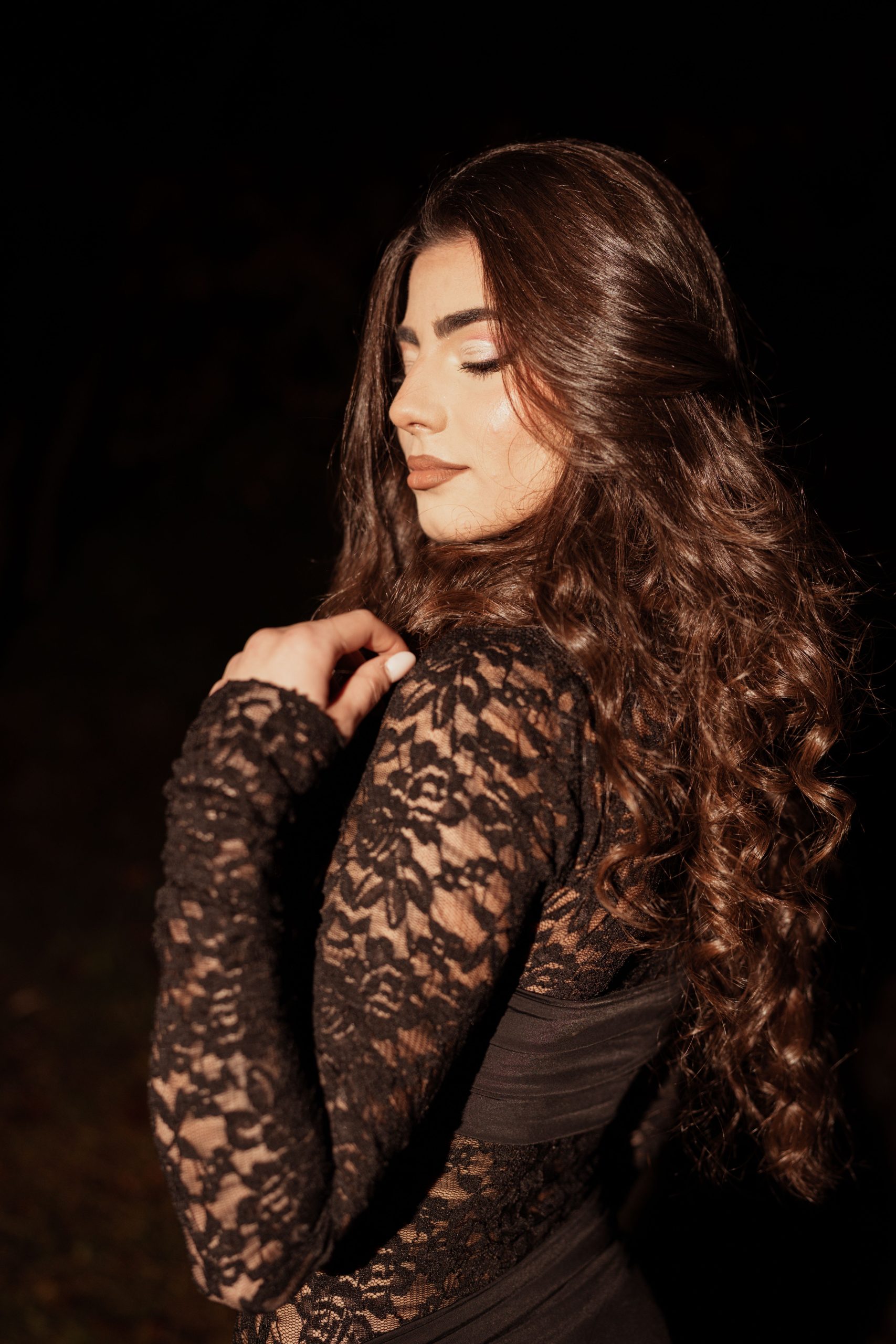 Stylish woman in black lace dress posing outdoors at night. Dramatic lighting highlights her elegant features.